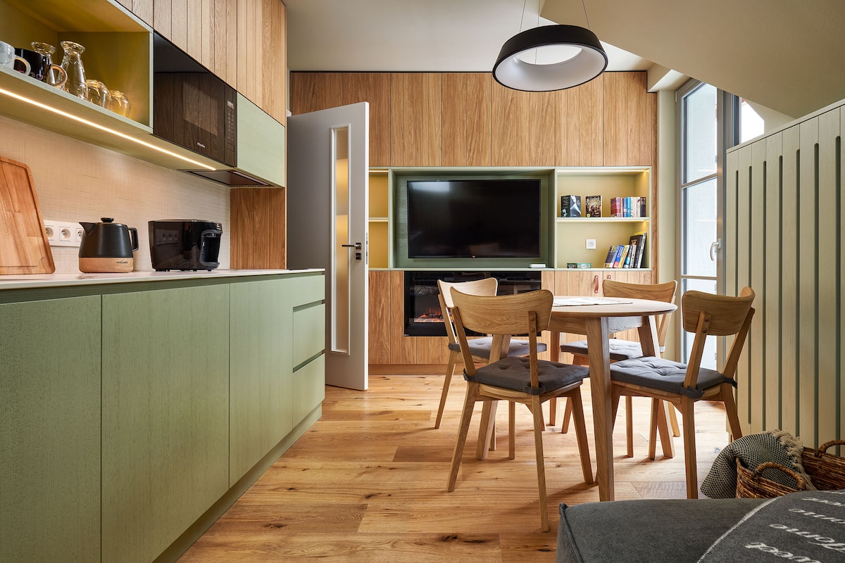 A modern kitchen and dining area features light wood cabinets paired with green accents. A small dining table with four wooden chairs is positioned under a circular pendant light. A flat-screen TV is mounted on the wall, complemented by shelves containing books.