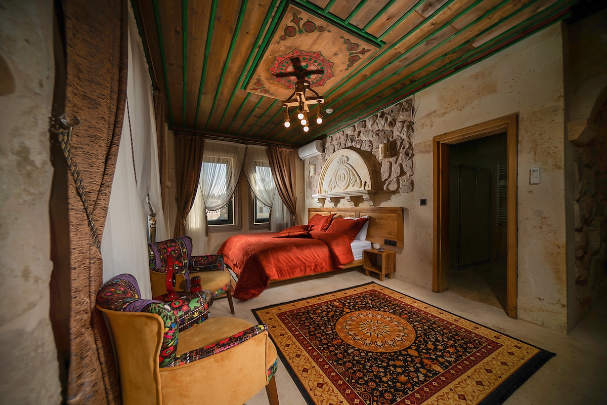 A spacious room is highlighted by an ornate ceiling and textured stone walls. A large bed with rich red bedding is complemented by two seating areas, featuring patterned chairs. Large windows are adorned with curtains, allowing natural light to brighten the space.