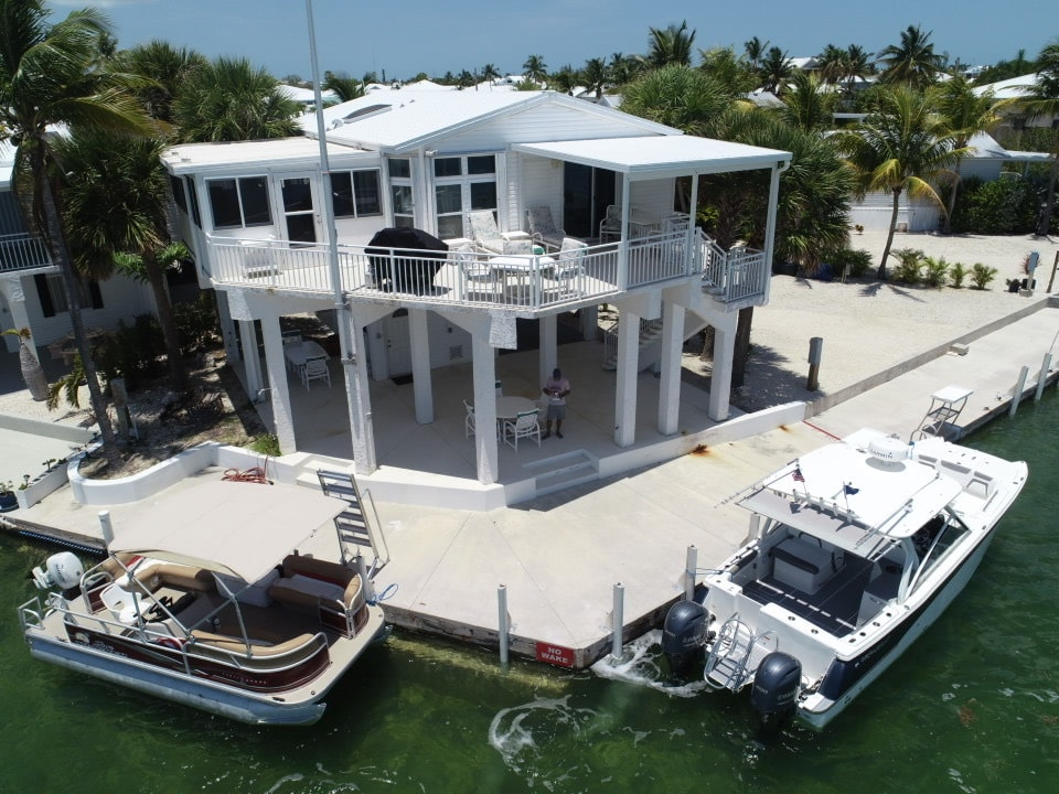 A waterfront property features a raised structure supported by pillars, with an outdoor seating area equipped with multiple chairs and tables. Two boats are docked alongside, and palm trees line the shore, contributing to a coastal ambiance.
