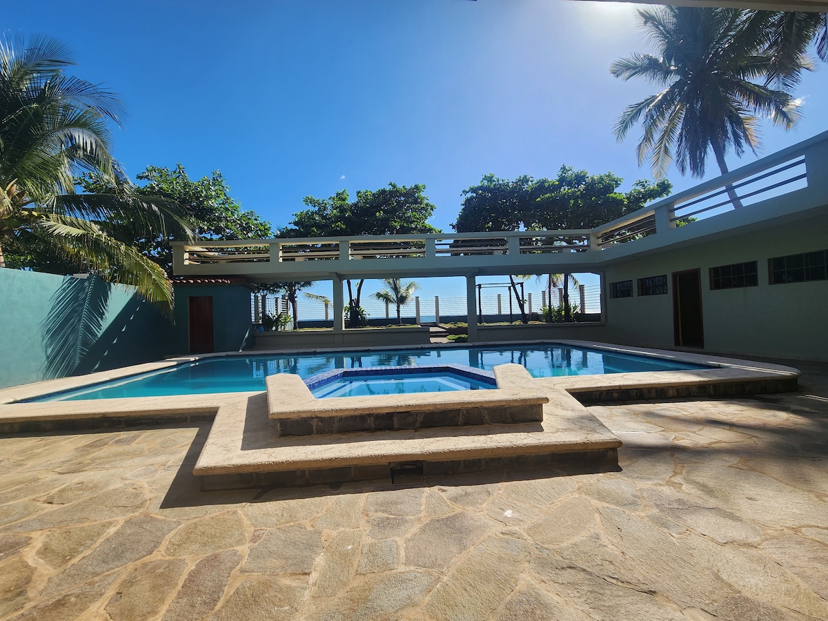 A spacious outdoor area features two swimming pools, one with a distinctive shape, surrounded by palm trees. A sunlit patio area can be seen in the background, providing a view of the ocean. The flooring is made of natural stone, creating a seamless transition to the pools.