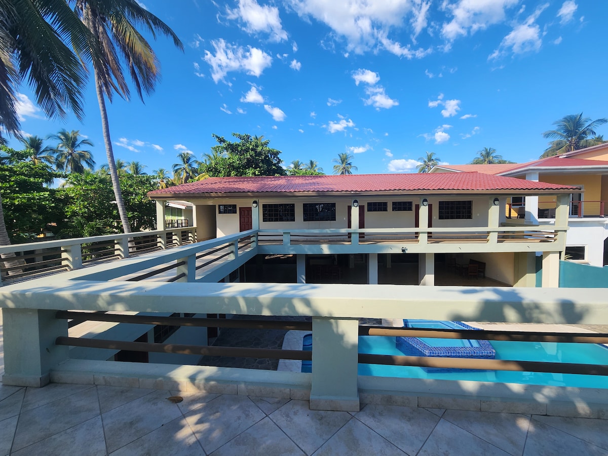 A view of the property features a two-story house with a red-tiled roof surrounded by palm trees. A spacious terrace overlooks a swimming pool, integrating outdoor space with the building's architecture. Bright blue sky and scattered clouds enhance the inviting tropical atmosphere.