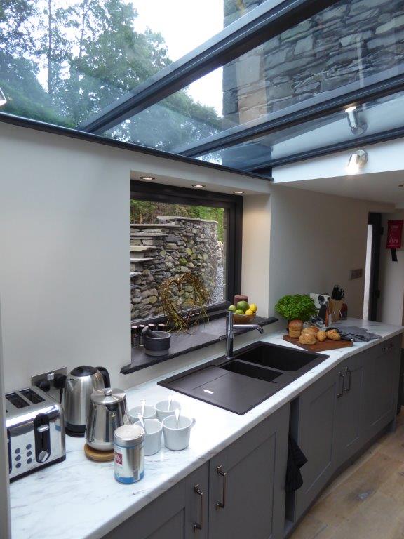 A modern kitchen features a sleek countertop with a sink, kettle, toaster, and coffee maker. Natural light floods in through large glass windows, illuminating a bowl of fruit and a fresh herb plant. Breads and pastries are neatly arranged on a wooden board.