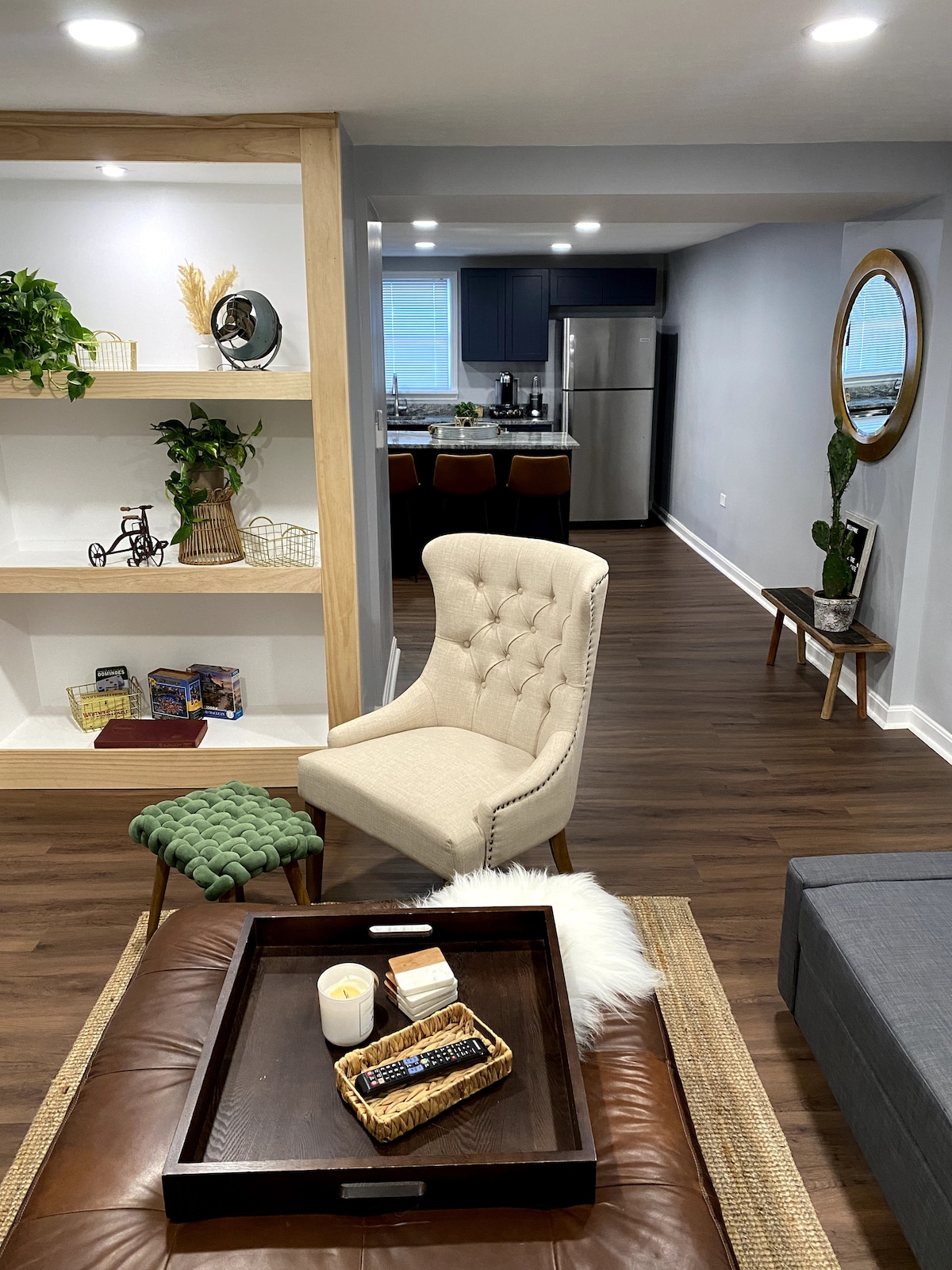 A cozy living area is highlighted by a plush armchair and a green ottoman. A wooden tray holds decorative items, while a built-in shelf displays plants and books. The kitchen is visible in the background, featuring modern appliances and a neutral color palette.
