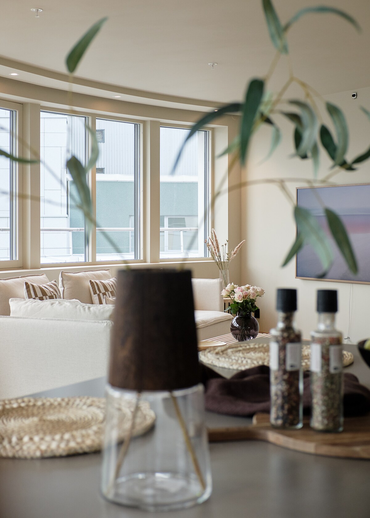 A well-lit living area with large windows allows natural light to filter in. In the foreground, a decorative glass container holds greenery. Nearby, spice jars and a wooden serving board rest on a table, creating an inviting atmosphere.