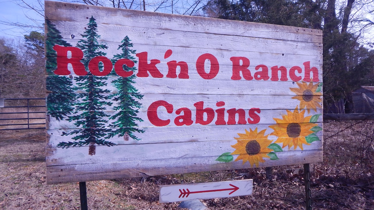 A rustic wooden sign announces 'Rock'n O Ranch Cabins' in bold red letters, accompanied by painted green trees and bright sunflowers. The sign is set against a natural backdrop of trees, offering a welcoming entrance to the cabins.