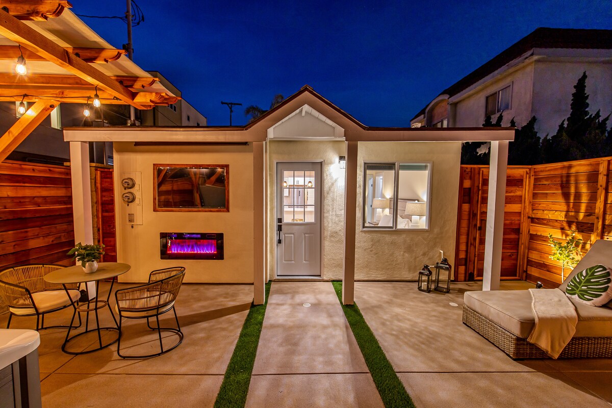 An outdoor area features a charming seating arrangement with two chairs and a table, illuminated by string lights. The wall-mounted electric fireplace creates a cozy ambiance, while the entrance to the villa is framed by a covered porch, inviting guests to enjoy the private space.