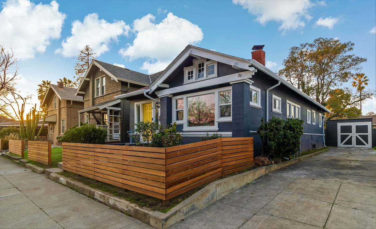 A single-family home is showcased with a welcoming blue exterior and decorative wooden fencing. A neatly landscaped yard is visible, featuring green plants and a gravel driveway. The garage is seen on the side, complementing the overall design of the residence.