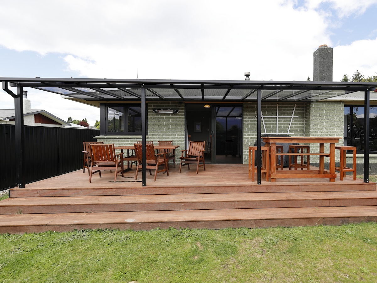 A large covered outdoor area features several wooden dining tables and chairs arranged on a wooden deck. The green brick house is visible in the background, with large windows that allow for natural light. The surrounding grass is well-maintained, enhancing the outdoor atmosphere.