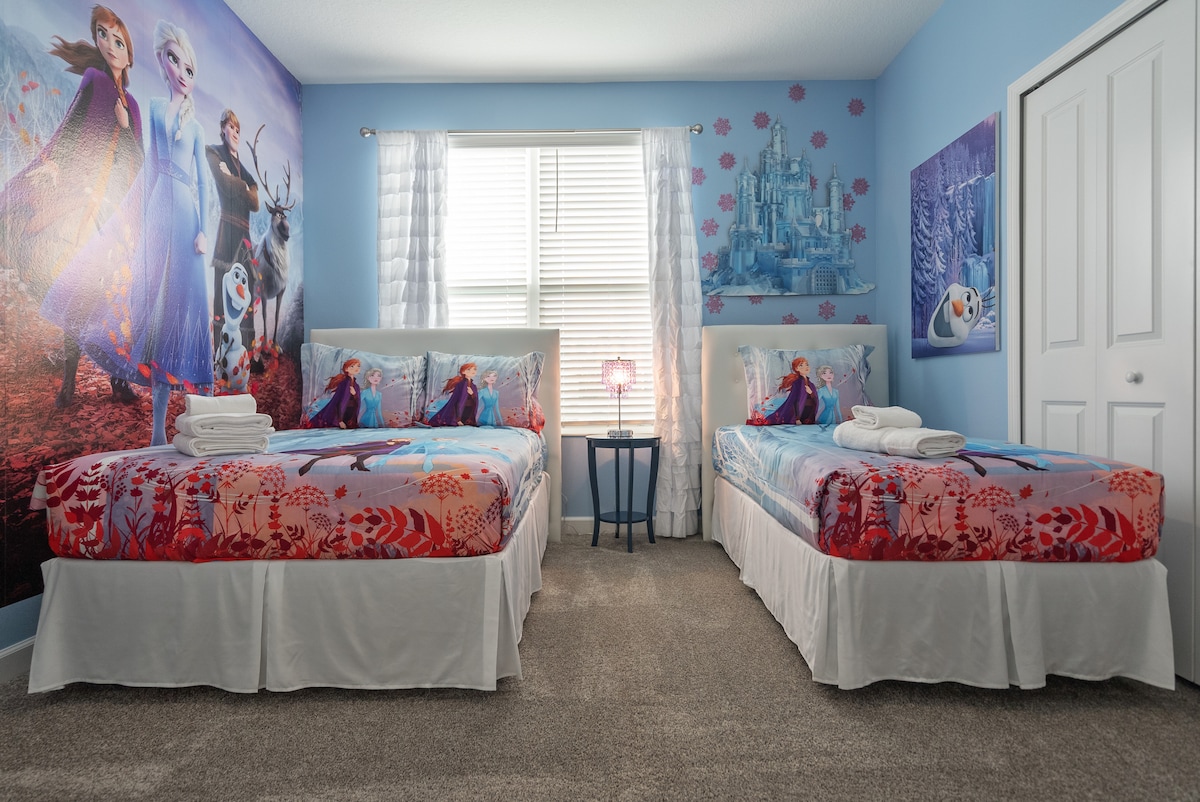 A children's bedroom features two spacious beds, each adorned with a colorful 'Frozen' themed bedspread. The walls are decorated with vibrant illustrations of familiar characters, and a small blue bedside table holds a lamp. Natural light streams through the window, complemented by sheer white curtains.