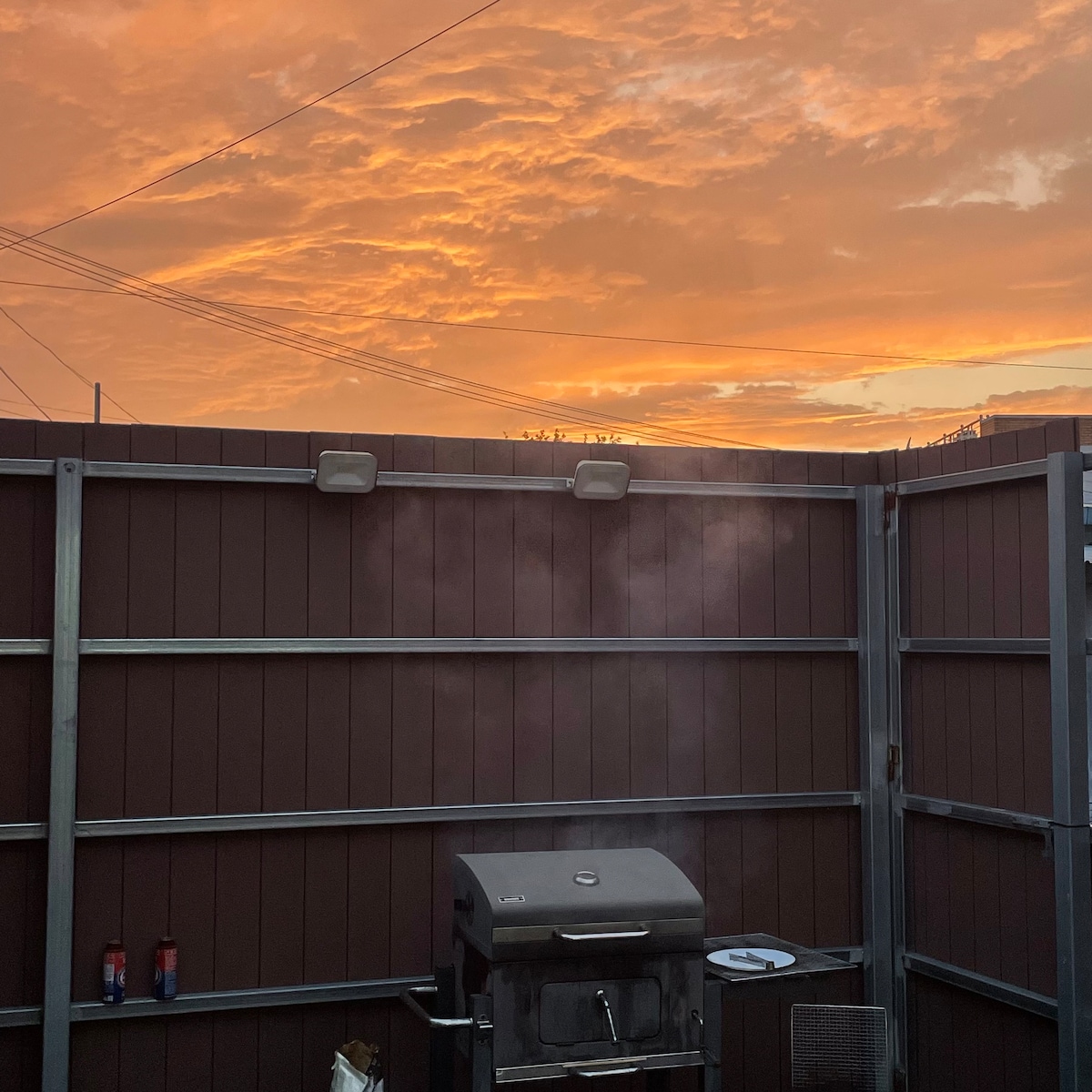 A BBQ grill is positioned against a wooden fence with a vivid sunset sky displaying orange and pink hues. The scene conveys a peaceful outdoor area, ideal for evening gatherings.