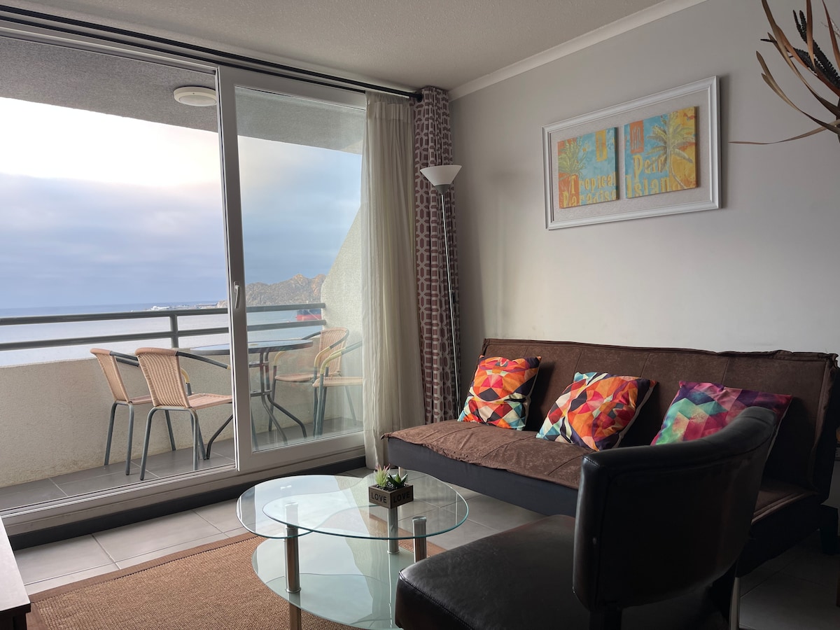 A living area is shown with a cozy sofa adorned with colorful cushions. A glass coffee table sits in front, and large sliding doors open to a balcony with a table and chairs, providing views of the ocean.