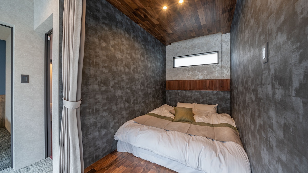 A cozy sleeping area is presented with a spacious bed featuring neutral bedding and a decorative cushion. The walls are finished in a textured dark grey, contrasting with warm wooden accents on the ceiling and floor. A small window allows natural light to illuminate the room.