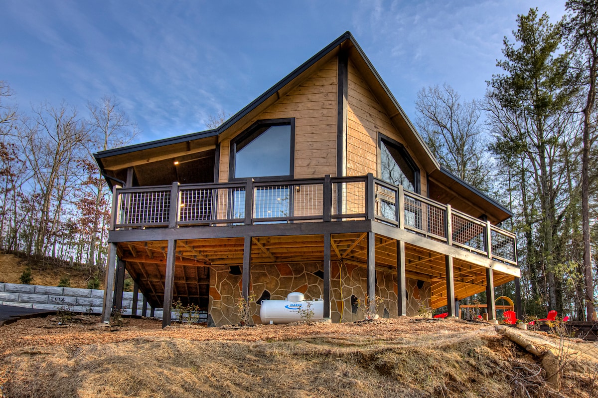 A new cabin is showcased, featuring a wooden exterior and large windows that provide ample natural light. The property is elevated on a stone foundation, with a spacious deck wrapping around the front. Red outdoor chairs are positioned on the lower level, surrounded by natural scenery.