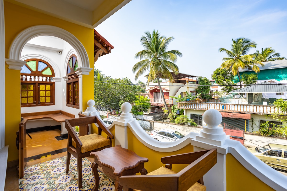A balcony is presented with two wooden chairs and a small table, offering a view of palm trees and neighboring houses. Bright sunlight illuminates the area, highlighting the colorful tiled floor and intricate architectural details of the balcony's railing.