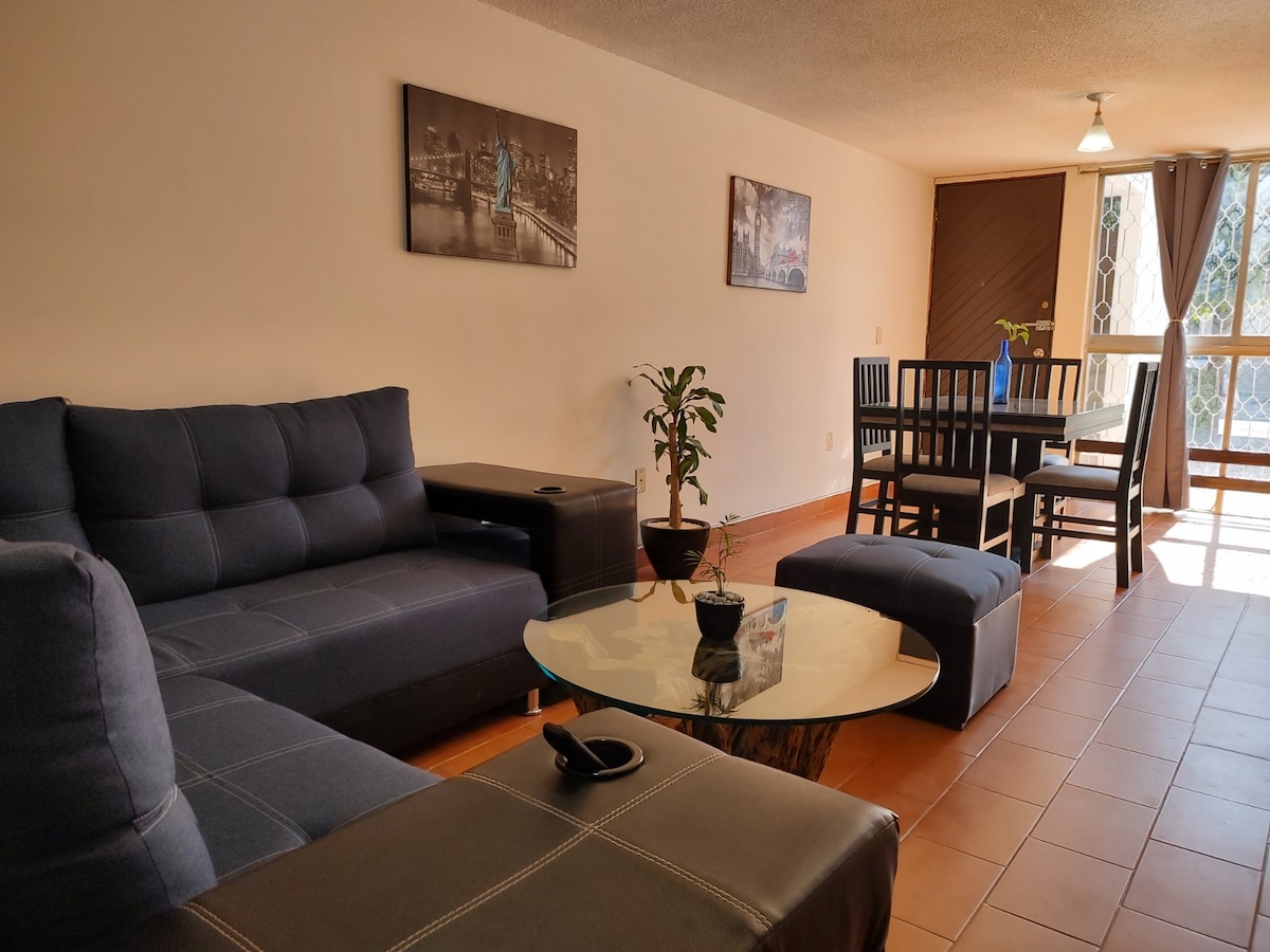 A cozy living area features a dark sectional sofa and a glass-top coffee table with a small potted plant. Natural light enters through large windows, illuminating a dining table surrounded by chairs, complemented by artwork on the walls.