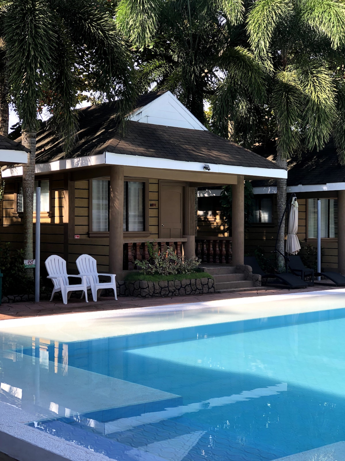 A charming cottage is framed by tall palm trees, with a clear blue pool in the foreground. Two white chairs are positioned to the side, inviting relaxation. The cottage features wooden siding, a set of steps leading to a porch, and large windows allowing natural light.