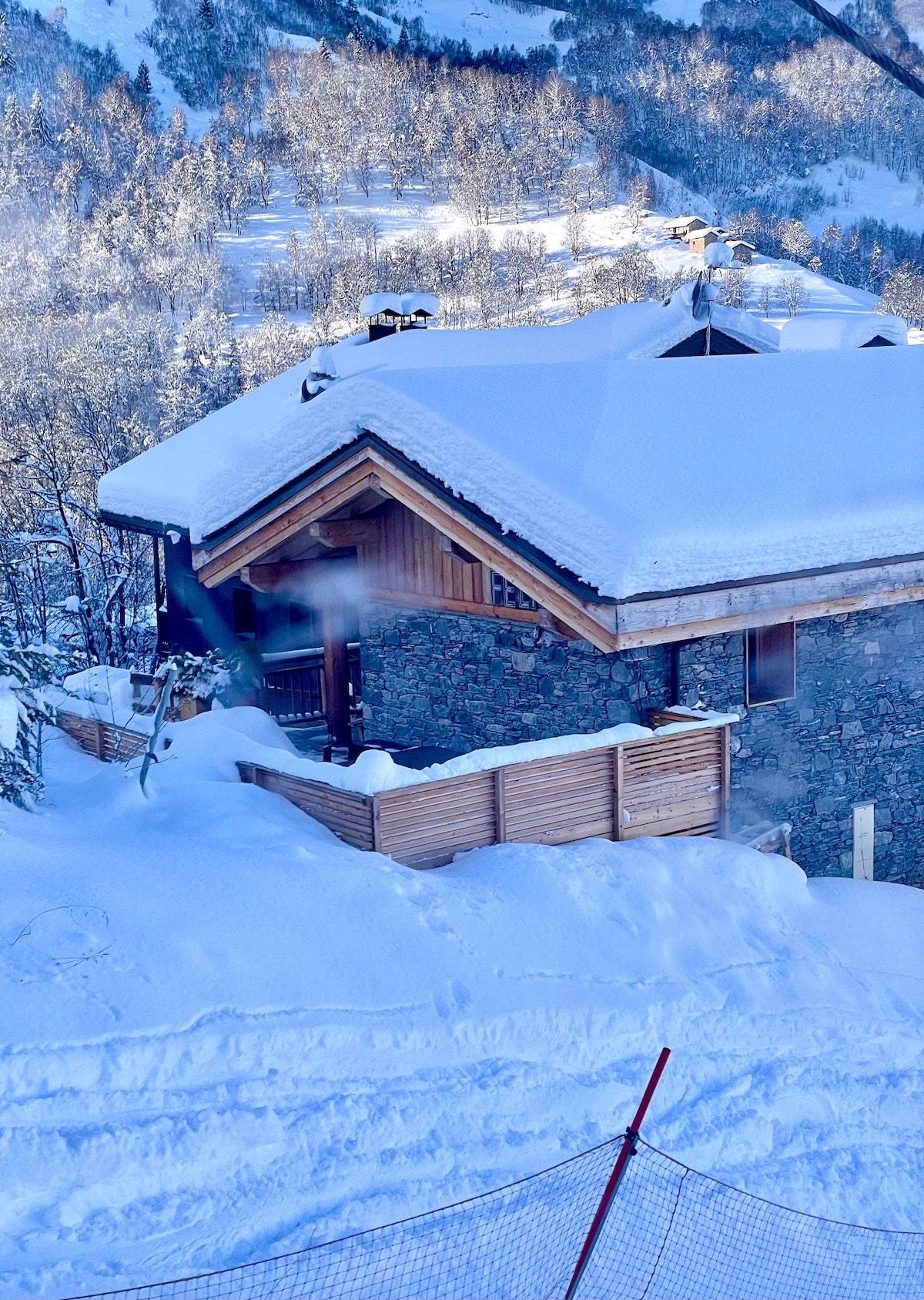 The chalet is nestled in a snowy landscape, featuring a wooden and stone facade. A sloped roof, covered in snow, complements the rustic charm. The private terrace, partially visible, is surrounded by snow, suggesting a serene winter retreat.