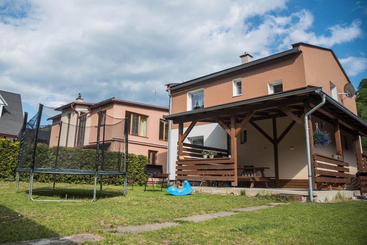 A spacious backyard features a wooden-covered patio with seating, providing an area for relaxation. Adjacent to the patio, a trampoline is visible on the well-maintained lawn. The surrounding greenery offers privacy, while neighboring houses are seen in the background.
