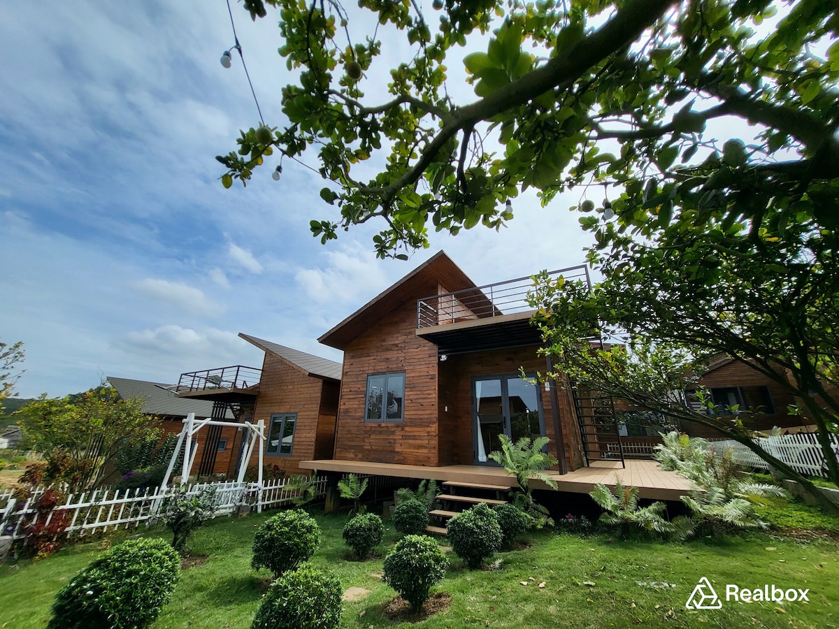A charming wooden house is nestled within a green landscape, featuring a large deck at the front. Surrounding foliage includes neatly trimmed bushes and vibrant plants, and a white picket fence encloses the property, enhancing its inviting appearance under a partly cloudy sky.