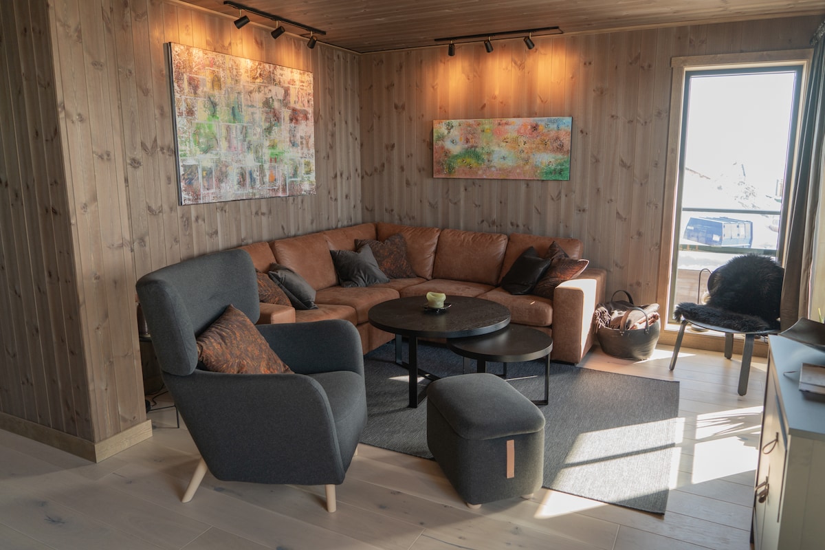 A modern living area features a large brown sectional sofa alongside a sleek round black coffee table. Natural light enters through a window, illuminating the wooden walls. Two colorful wall art pieces add visual interest, complemented by a soft grey chair and footrest.