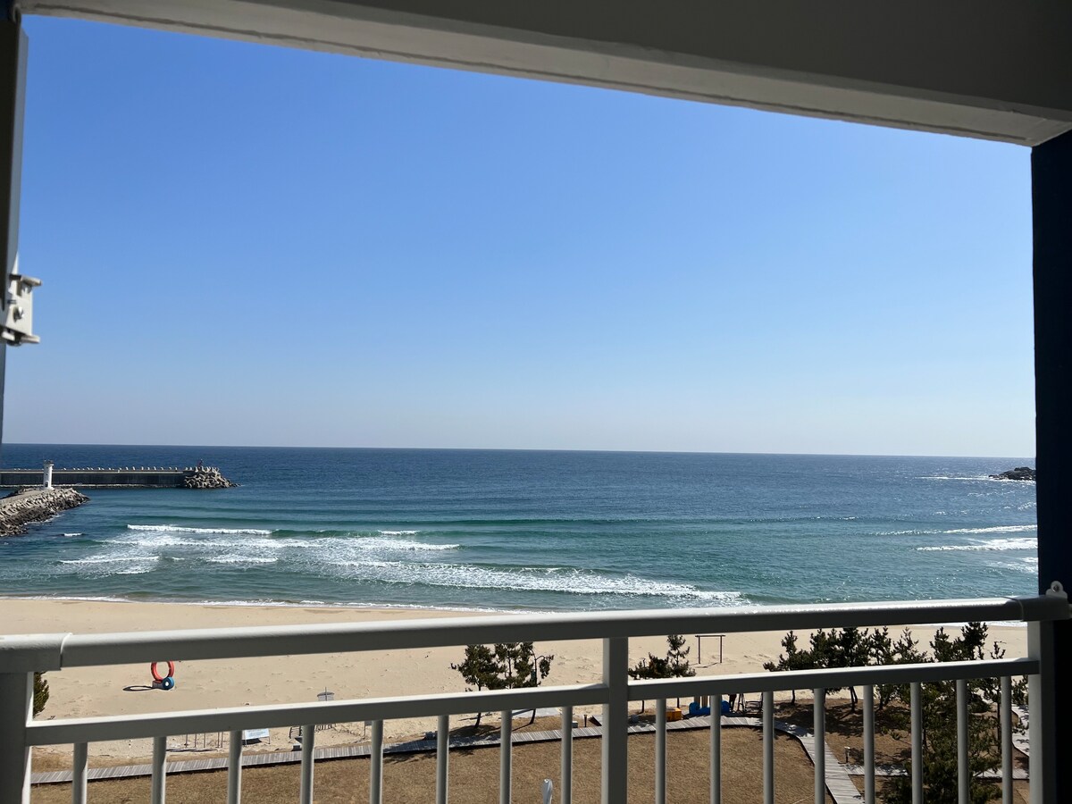 A view of the ocean is framed by a balcony railing. Gentle waves crash onto a sandy beach under a clear blue sky, with a distant pier visible along the shoreline. Nearby, hints of greenery and a glimpse of beach furniture can be spotted, contributing to a serene coastal atmosphere.