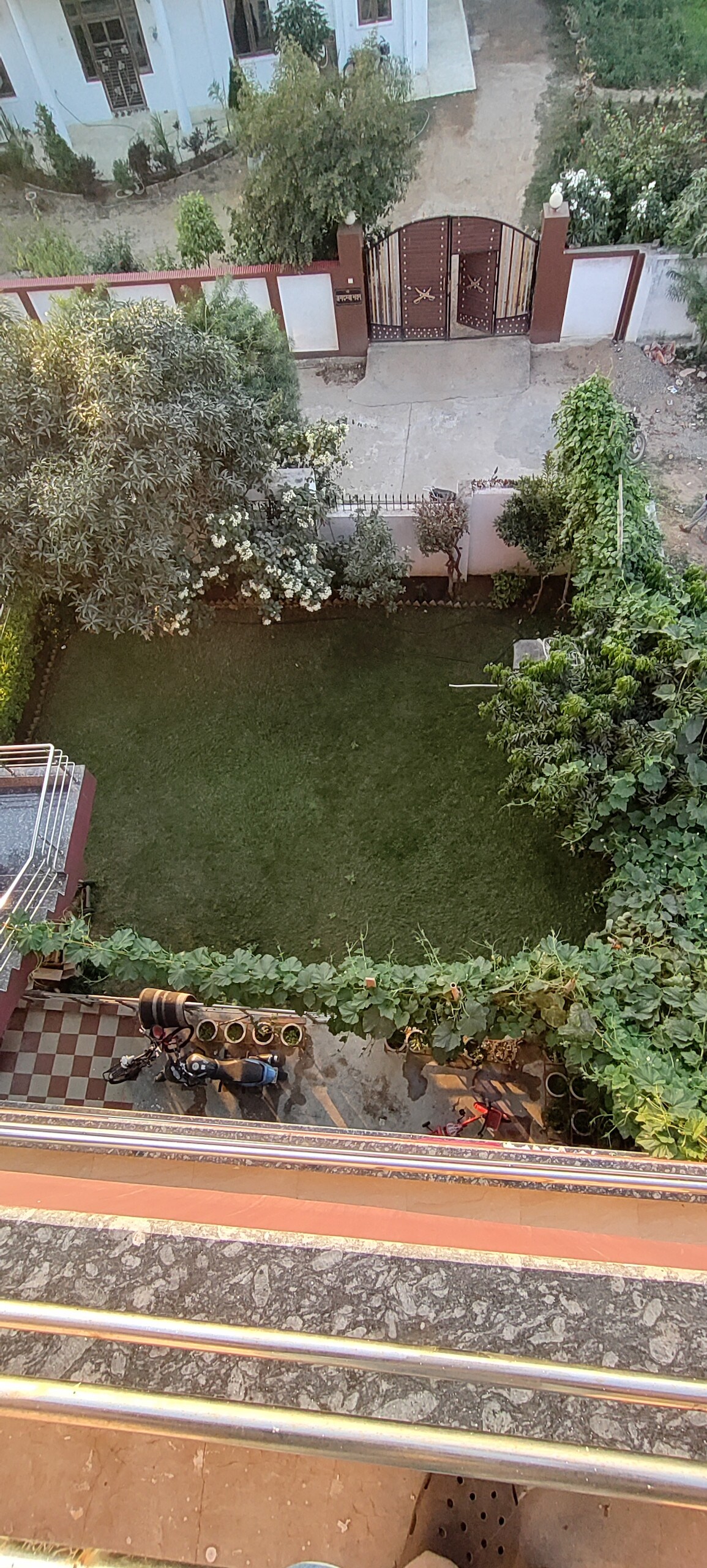 A well-maintained garden is visible from the balcony, featuring lush green grass and various plants. A path leads through the greenery, with decorative elements such as pots arranged along the edges. The entrance gate and surrounding fence provide a sense of privacy.
