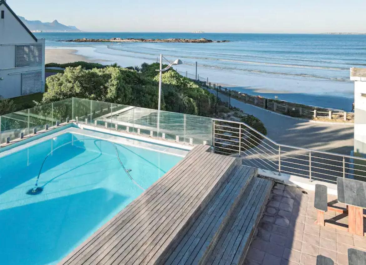 An outdoor pool area features a spacious wooden deck with steps leading down to the water. The pool is viewed alongside the ocean, which stretches into the distance, providing a clear view of the shoreline and surrounding greenery.