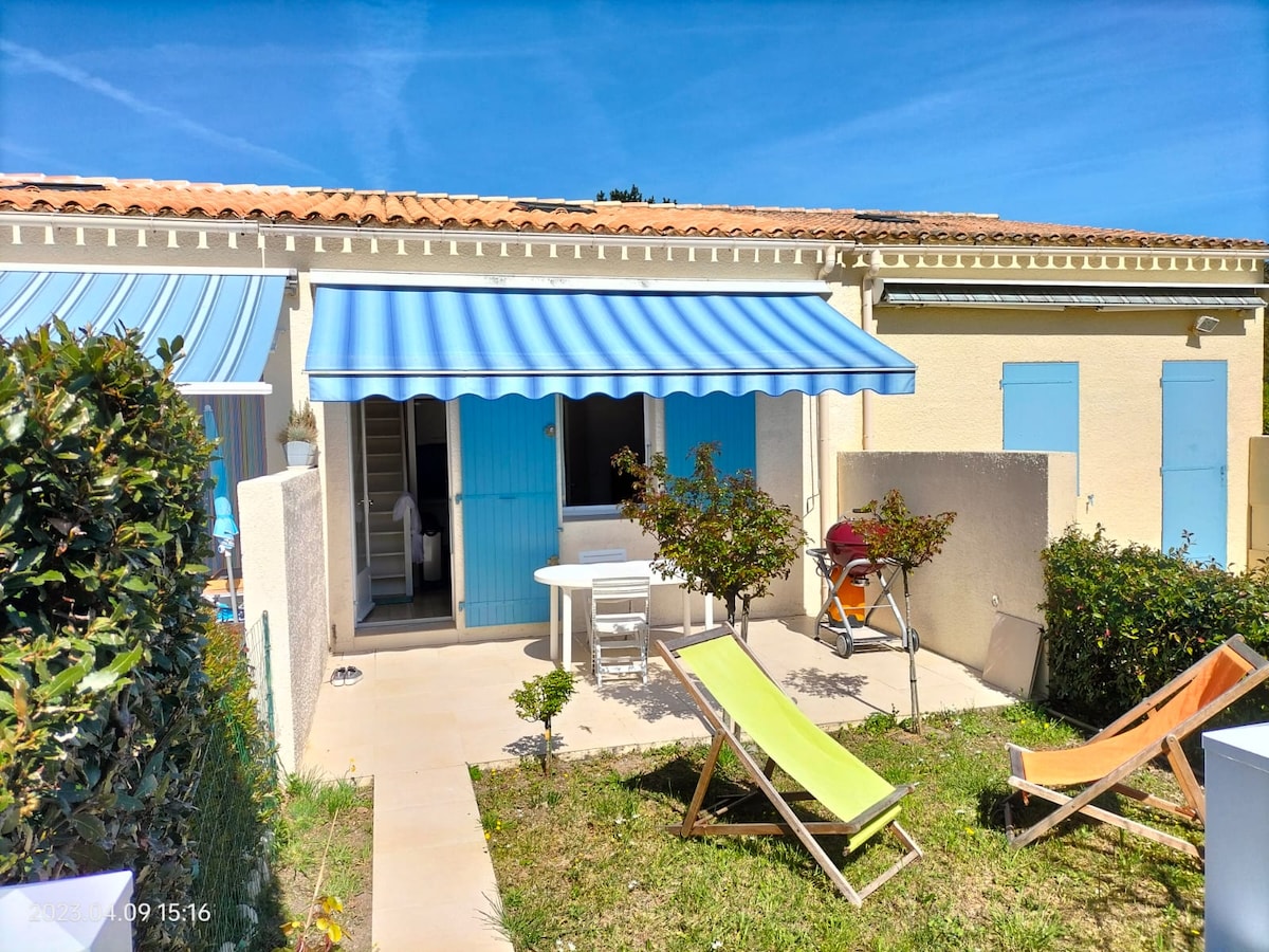 The exterior features a charming terrace with a blue and white striped awning. A small garden is visible with two sun loungers and a dining table set. A vibrant green lawn surrounds the area, framed by low hedges and a nearby wall.
