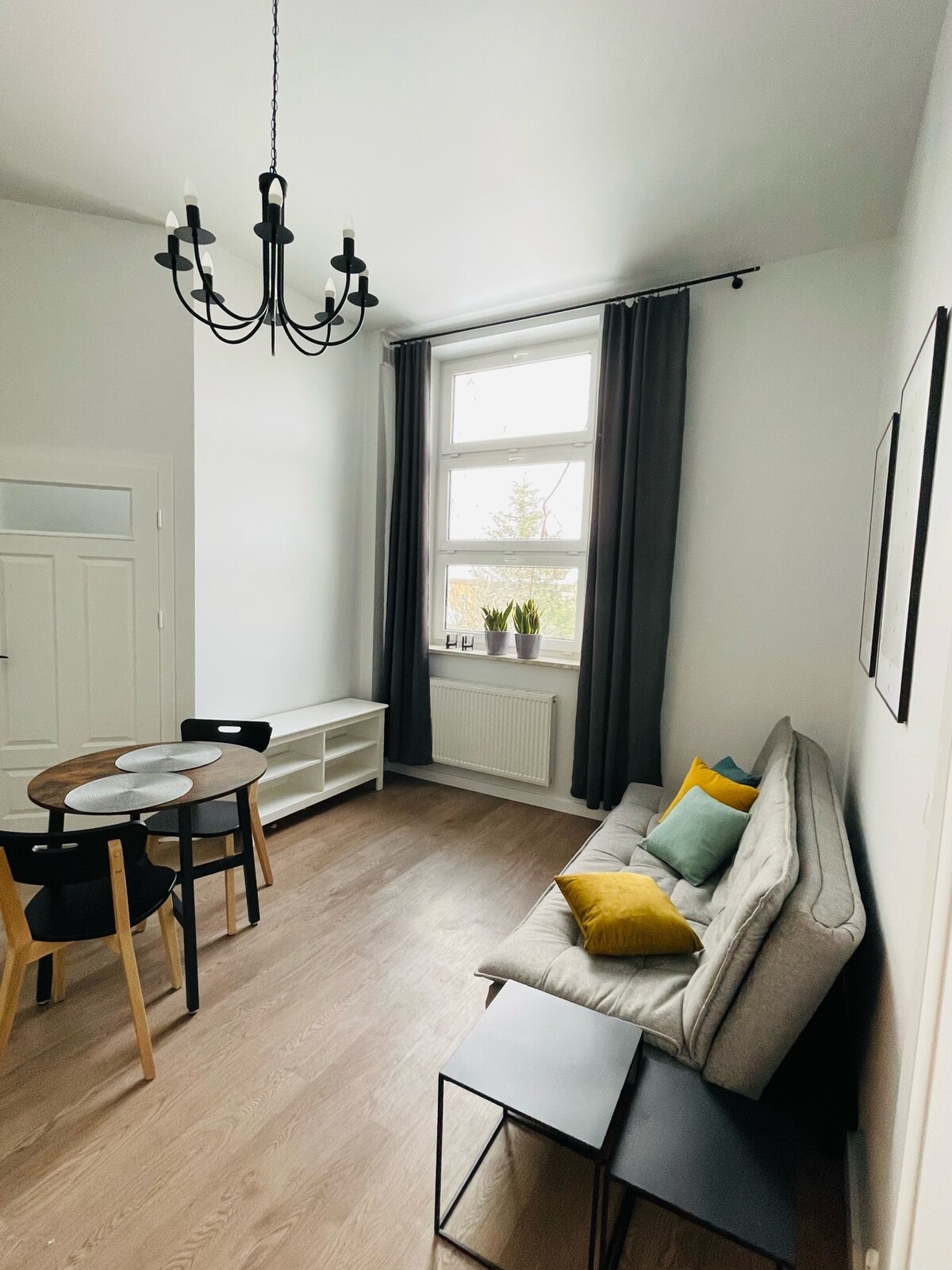 A light-filled living area features a cozy gray sofa adorned with colorful cushions. A round dining table with black chairs is positioned nearby, while a small black side table complements the space. Tall windows with dark curtains provide natural light, enhancing the inviting atmosphere.
