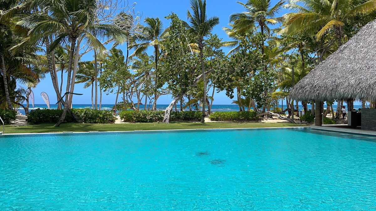 An expansive infinity pool reflects the clear blue sky, surrounded by lush palm trees and open green spaces. The beach is visible in the background, providing a serene coastal backdrop.