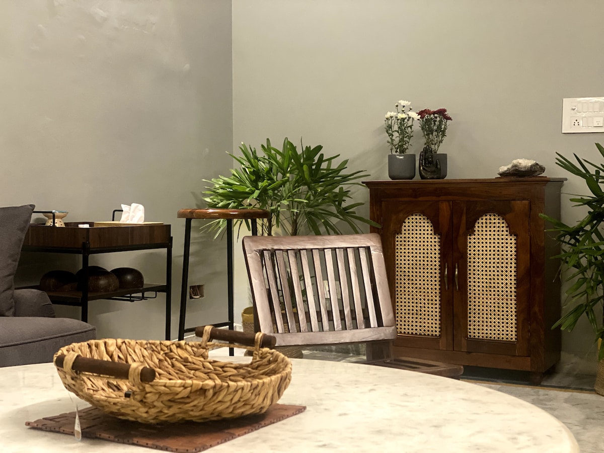 A cozy living space showcases a blend of modern and traditional decor. A wooden cabinet with intricate doors stands beside potted plants. A round marble table features a woven basket, while a comfortable chair and side table complete the inviting arrangement.
