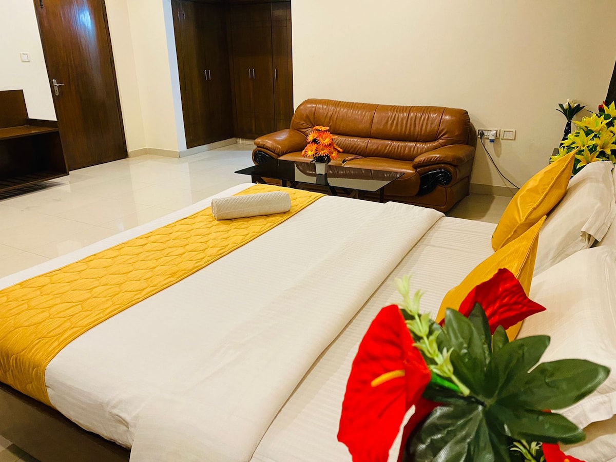 A spacious sleeping area features a large bed adorned with a light yellow blanket and a rolled towel. A brown leather sofa is positioned nearby, complemented by potted plants with vibrant red flowers, adding a touch of color to the room.