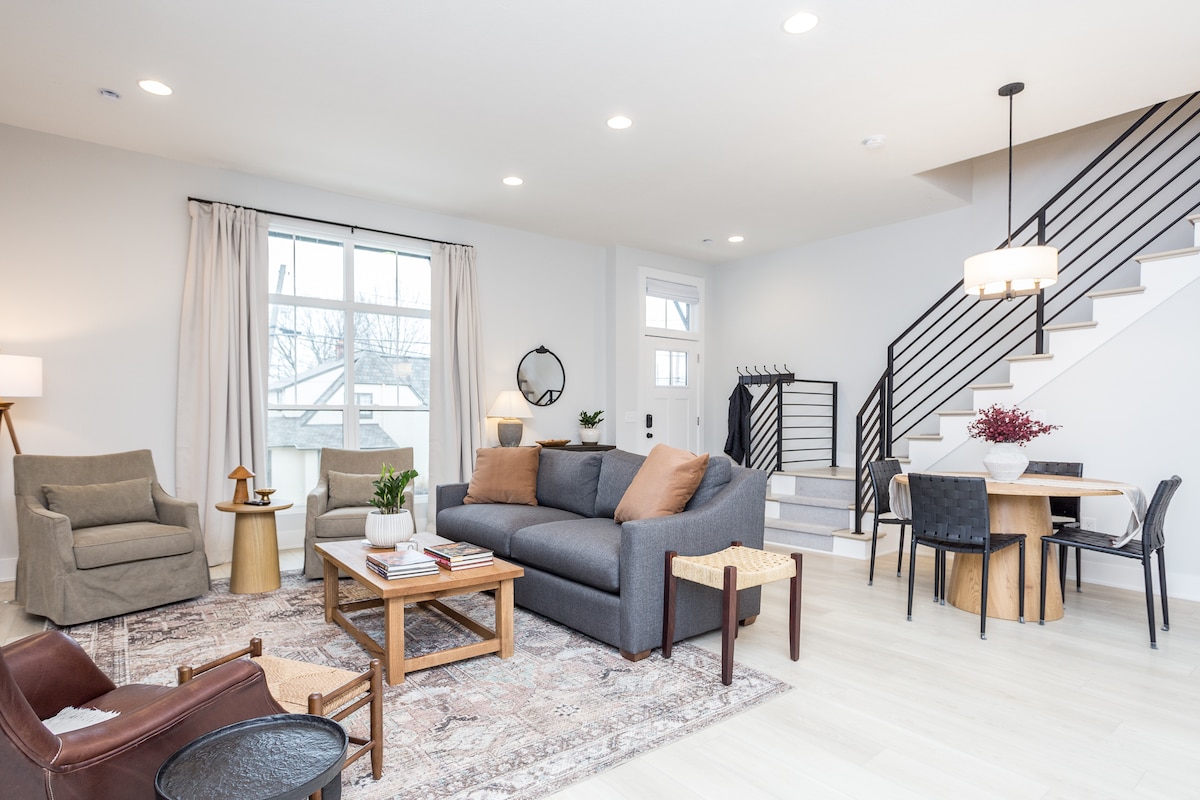 An open concept living area features modern furnishings, including a spacious gray sofa and two accent chairs. Natural light enters through a large window, illuminating a wooden coffee table and a dining table set for four. A staircase leads to the upper level.