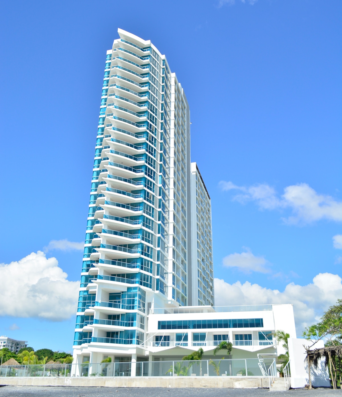 A tall, modern building with a sleek design is depicted against a clear blue sky. The facade features large glass windows and multiple balconies, highlighting the contemporary architecture. The surrounding area is landscaped with palm trees and pathways.