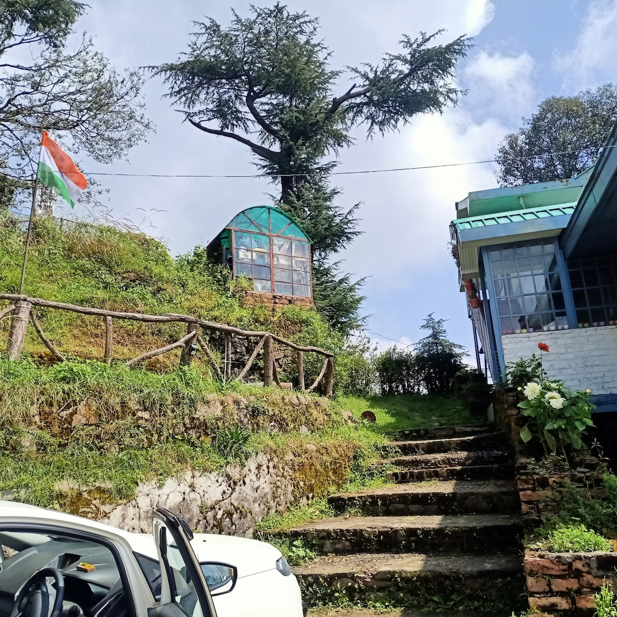 A steep, grassy hill adorned with an Indian flag features a quaint greenhouse structure at its peak. Stone steps lead up the hill, flanked by greenery and rustic wooden railings. Buildings in the vicinity include a blue-roofed house and a white structure.