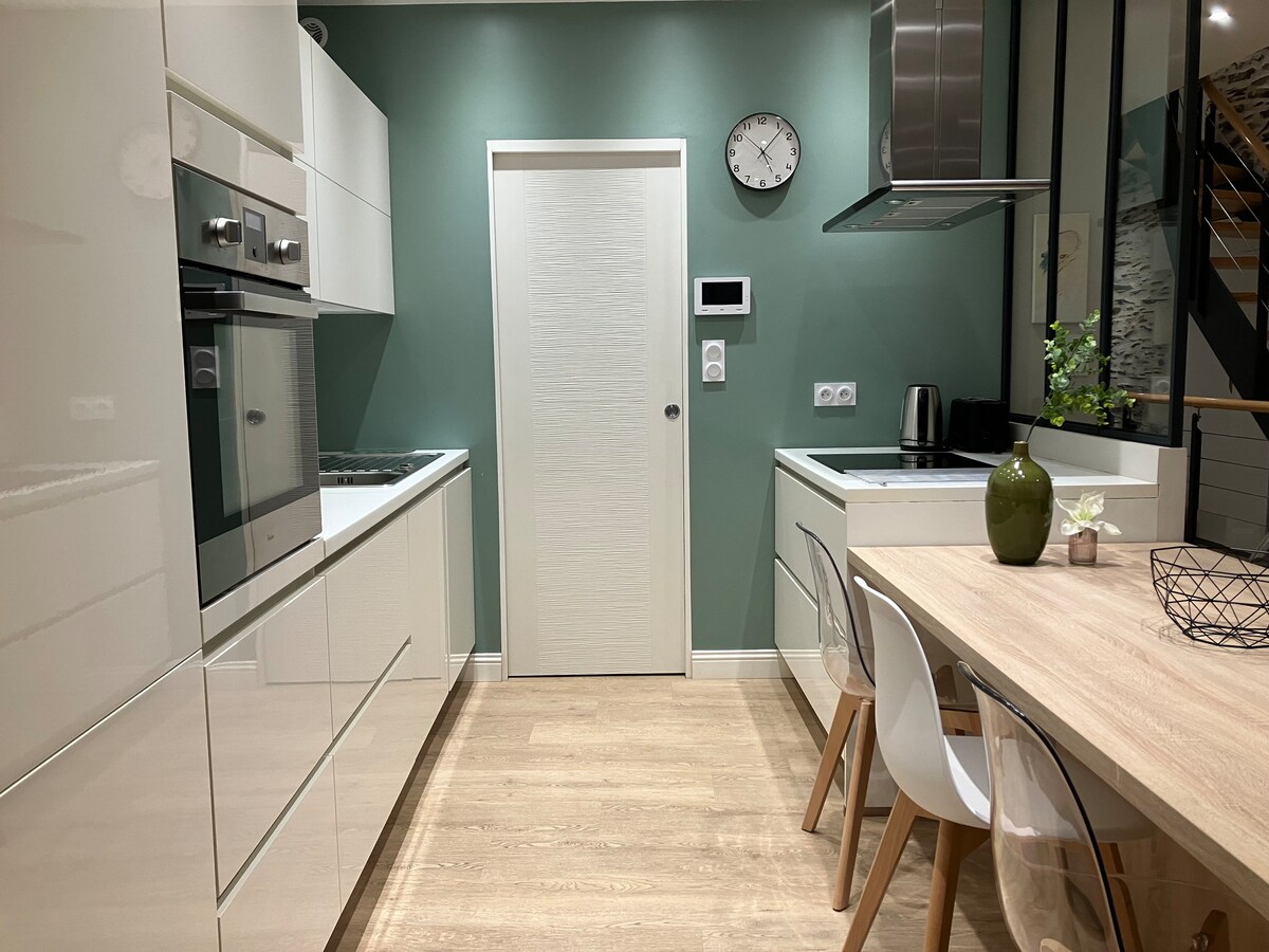 A modern kitchen is presented with sleek white cabinetry and a wooden dining table. The space features integrated appliances and a muted green accent wall. A clock is mounted on the wall, providing a functional touch. Natural light fills the area, creating a bright and airy atmosphere.
