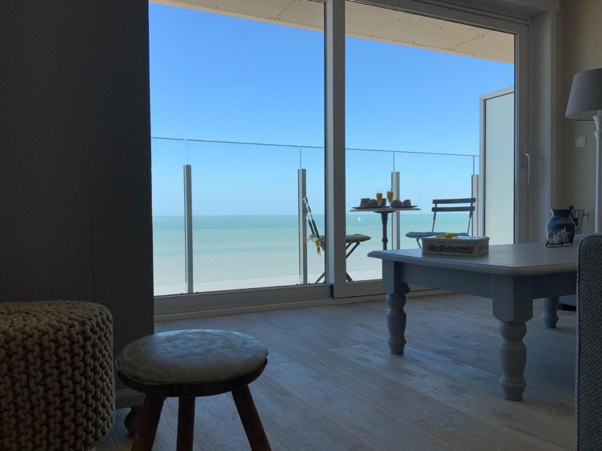 A bright room features large glass doors that open to a balcony with a table and two chairs, offering views of the calm sea and clear sky. A cozy ottoman and a light coffee table are present, creating a welcoming space for relaxation.
