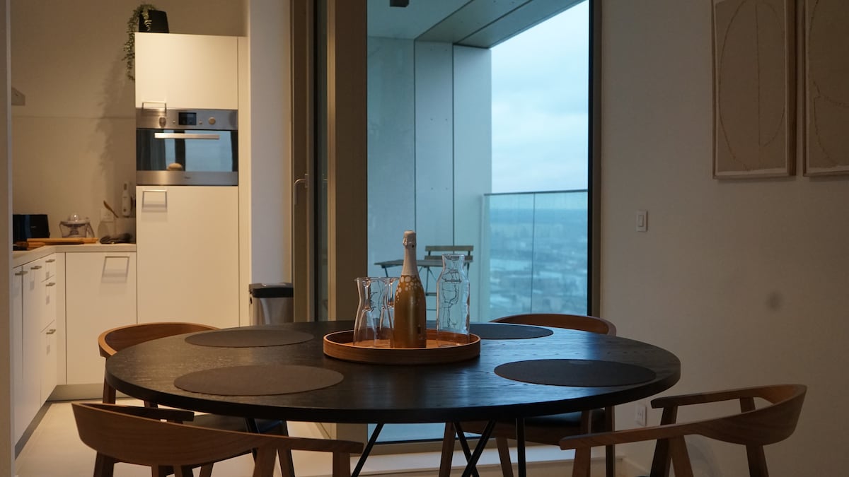 A round dining table with wooden chairs is positioned near large glass windows that offer expansive views. The minimalist kitchen features built-in appliances and a light color scheme. A wooden tray with several glass bottles is centered on the table, inviting shared meals.