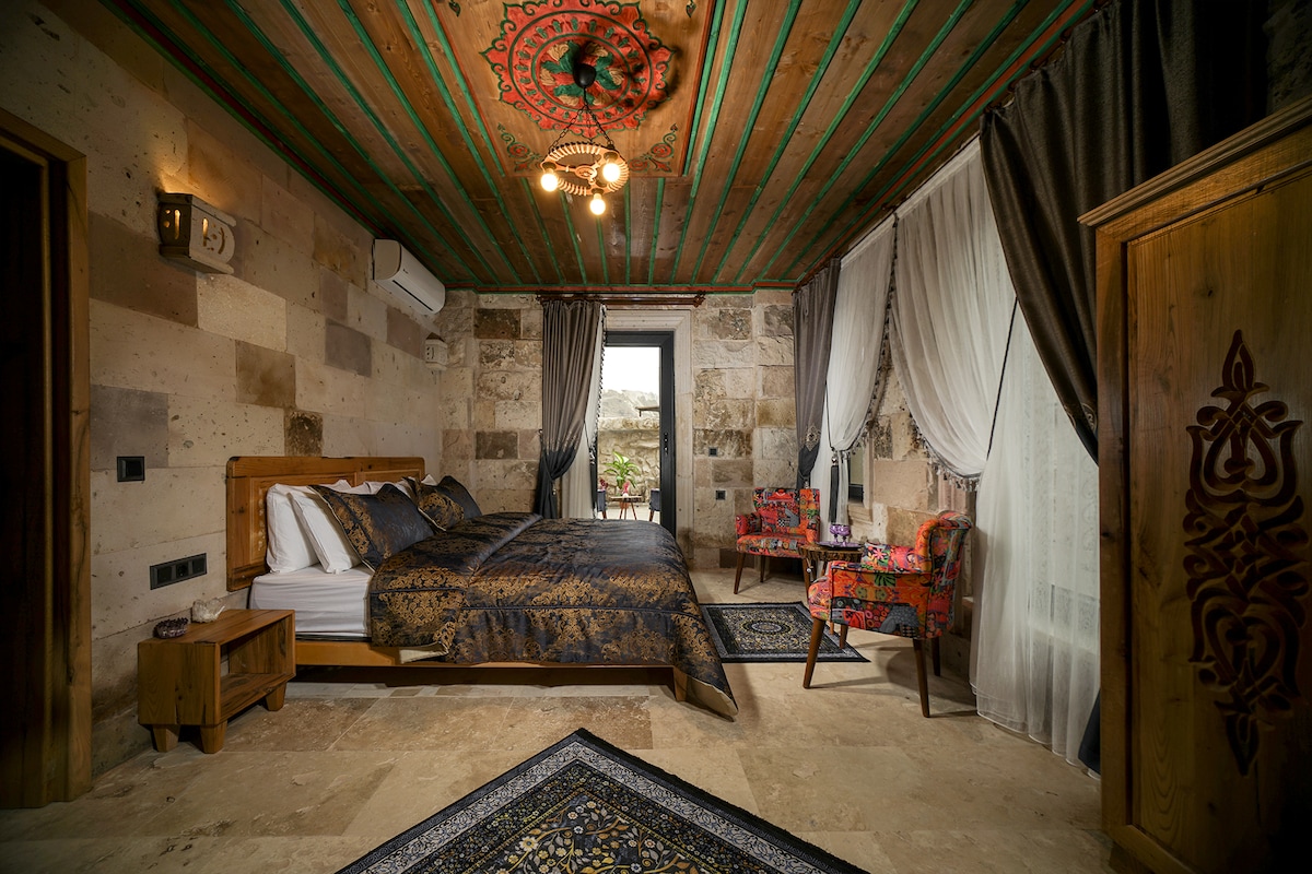 A deluxe studio suite features a spacious bed with intricate bedding and decorative pillows. Soft natural light filters through sheer and dark curtains, illuminating the stone walls and wooden accents. A colorful seating area complements the room, and a patterned rug enhances the inviting décor.