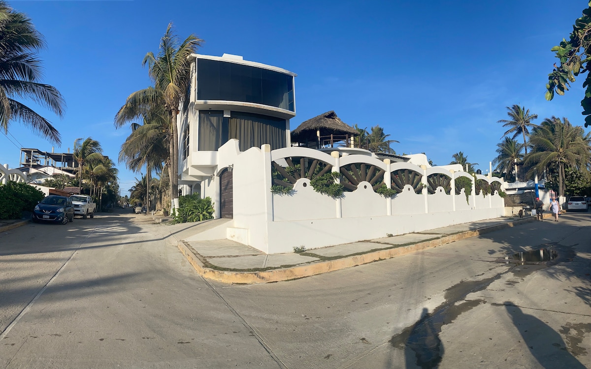 A modern, two-story building is set against a clear blue sky, surrounded by palm trees. The white facade is complemented by a curved wall at the entrance. The quiet street is lined with greenery, highlighting the tropical setting near the beach.