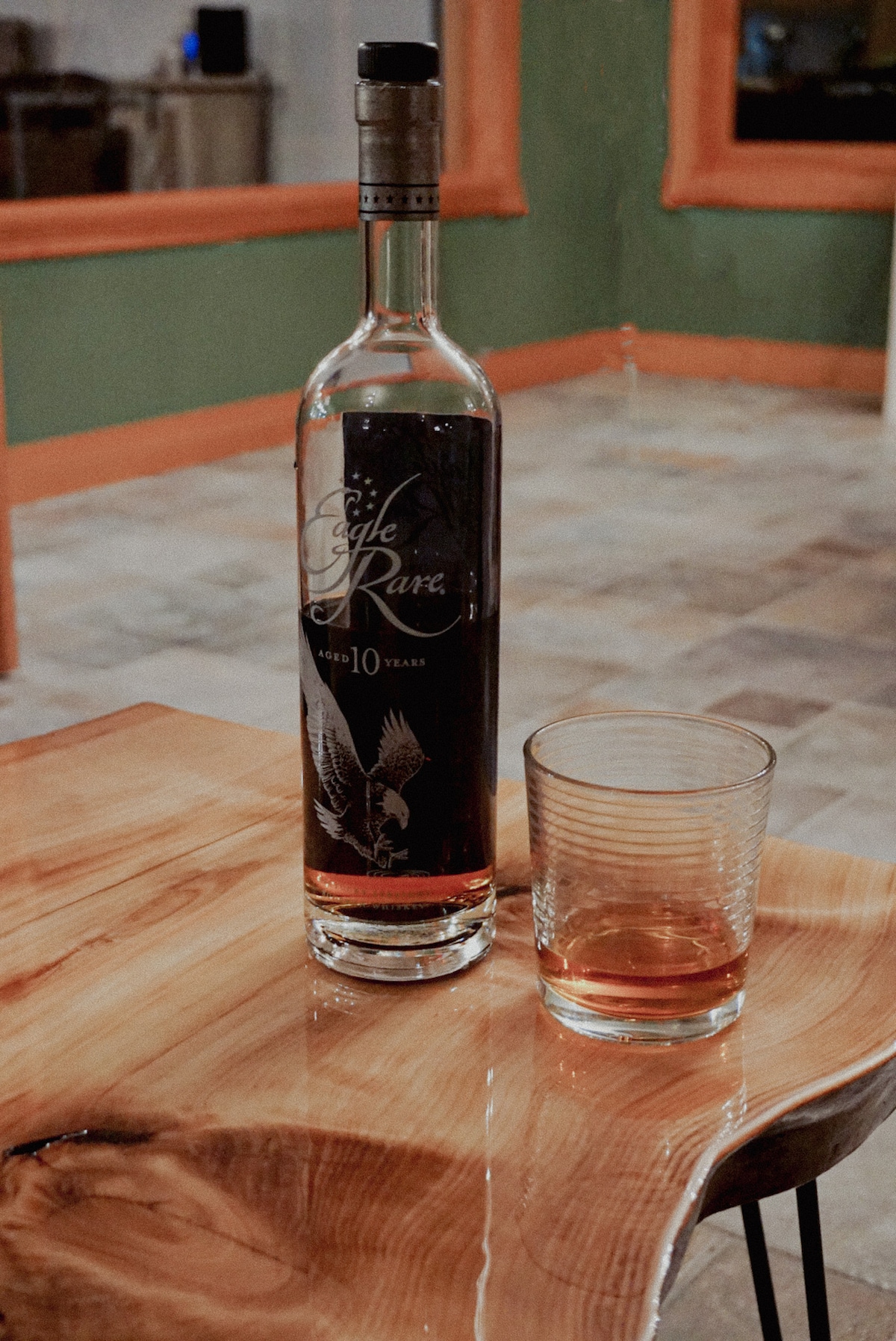 A bottle of Eagle Rare 10-Year bourbon stands on a wooden table with a smooth surface. Next to it, a glass filled with bourbon reflects the light, indicating a warm amber color. The setting features a softly lit background with neutral tones.