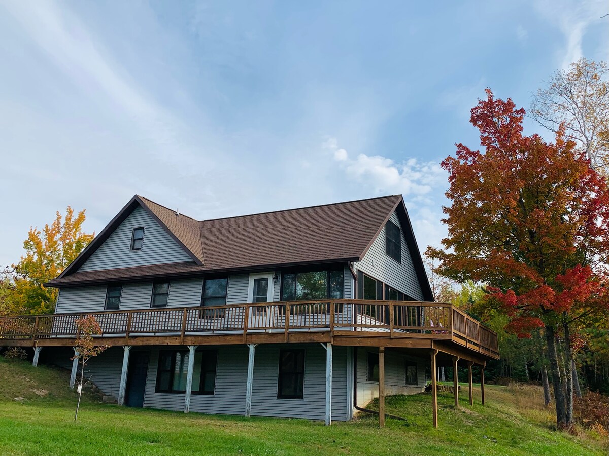 The exterior of a spacious cabin is displayed, featuring a wrap-around porch and large windows. Surrounded by greenery and autumn-colored trees, the structure has a steeply pitched roof and is set on a gently sloping lawn.