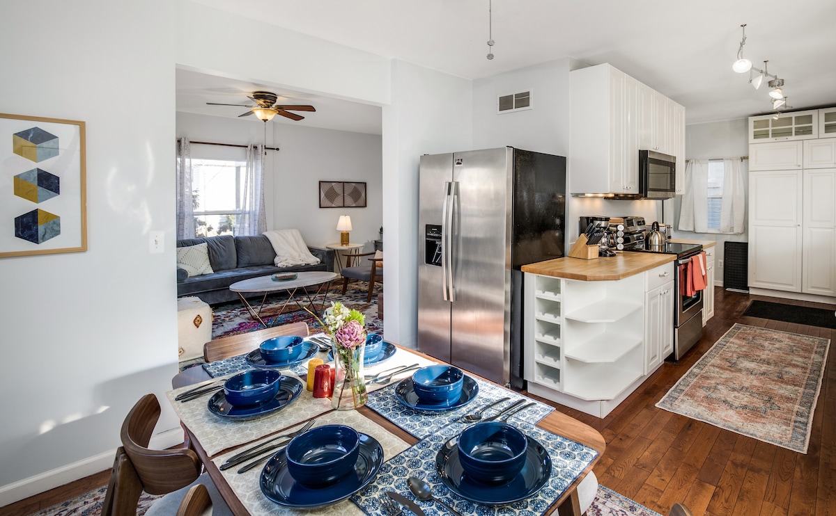 A well-appointed kitchen and dining area is showcased, featuring modern stainless steel appliances and a spacious wooden countertop. A dining table is set for four with blue plates, complemented by decorative table runners and a vase of flowers. The adjoining living area is partially visible.