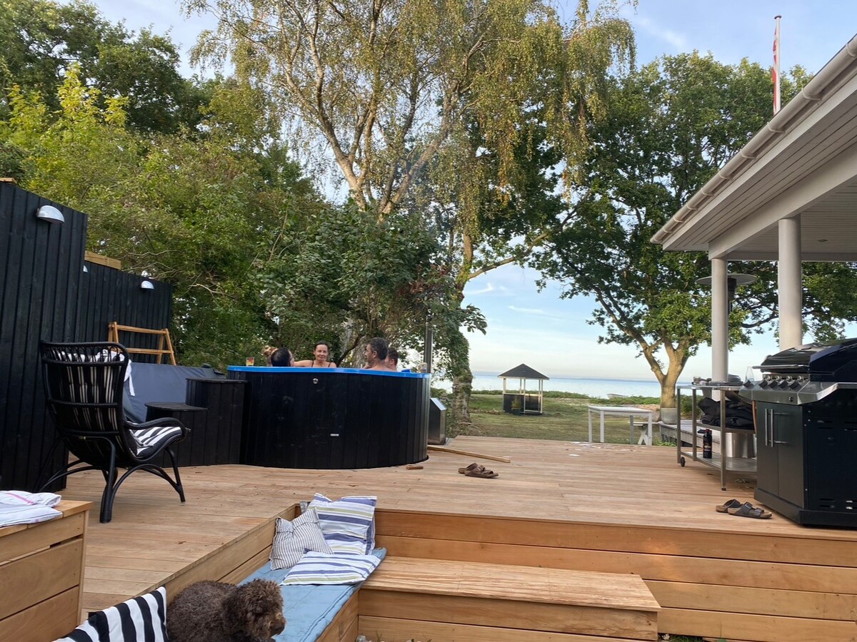 An outdoor deck area features a circular spa surrounded by lush greenery and a serene view of the coastline. Comfortable seating is available, with a barbecue station in the corner. The space invites relaxation and outdoor gatherings, complemented by a gentle breeze.