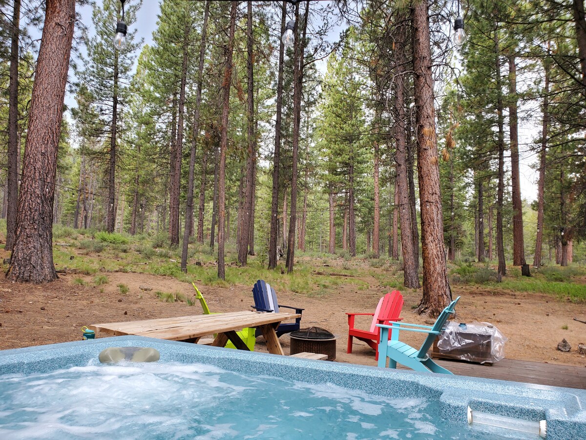 A serene backyard setting is visible with a hot tub bubbling next to a wooden picnic table. Colorful Adirondack chairs in red, blue, and purple are scattered around, surrounded by towering pine trees and a natural forest floor.