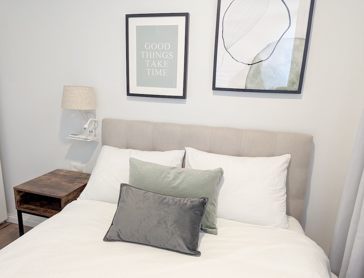 A cozy sleeping area features a modern upholstered headboard with plush white bedding. Two decorative pillows enhance the bed's design. Above the bed, two framed artworks add a touch of style, while a small bedside table holds a lamp for soft illumination.