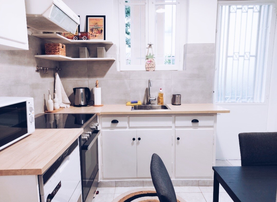 The kitchen features modern appliances, including a microwave and oven. A light-colored countertop provides workspace next to a sink. Shelves hold decorative items, and a small table with chairs is positioned nearby. Natural light enters through a window, brightening the space.