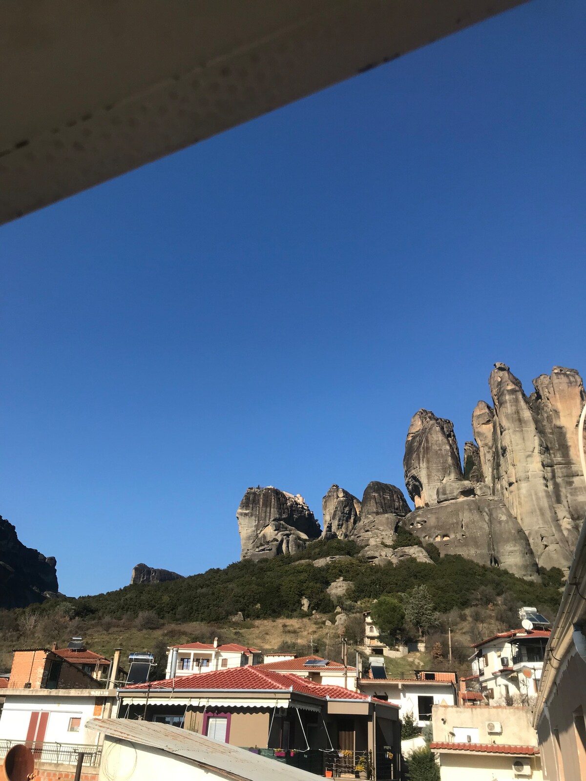 Majestic rock formations rise against a clear blue sky, creating a striking backdrop to the cozy village below. The rooftops of charming homes with warm tones are visible, nestled among green hills that add to the serene landscape.