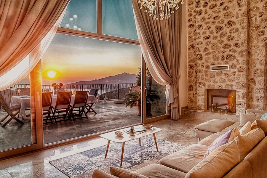 The living area features large glass doors that open to a terrace, offering a stunning view of the sunset over the mountains. Soft textured curtains frame the windows. A cozy sofa and a simple coffee table complement the inviting space, which is enhanced by natural light.
