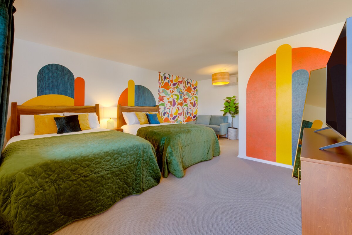 A vibrant guest room is highlighted by two spacious beds dressed in rich green covers. Colorful wall murals add artistic flair, while a plant and modern lamp contribute to the decor. A television is positioned on a wooden cabinet, offering entertainment options.